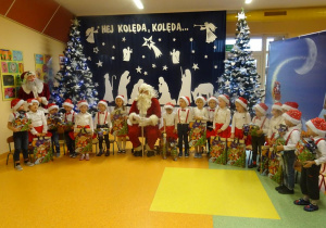 Grupa dzieci wraz z Mikołajem siedzi na krzesełkach, w tle świąteczna dekoracja.